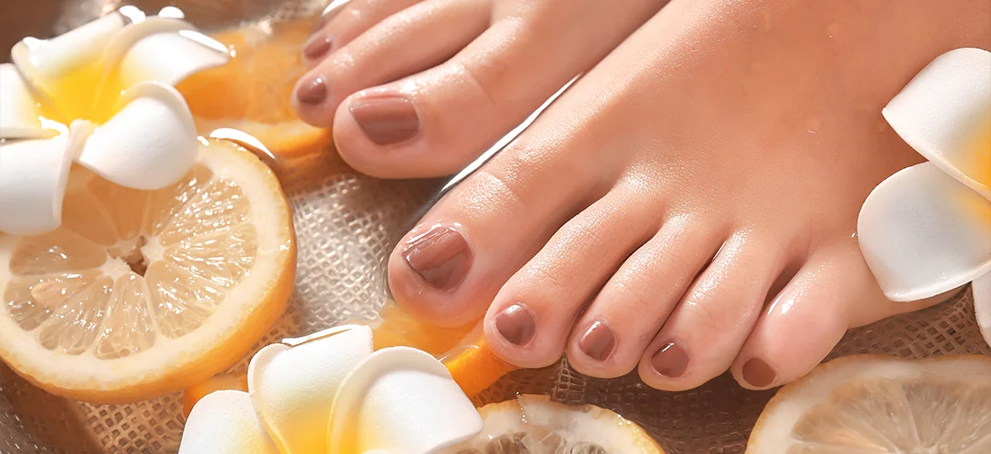 Client's feet resting in a clean pedicure soak with citrus slices and frangipani flowers, toenails painted mauve